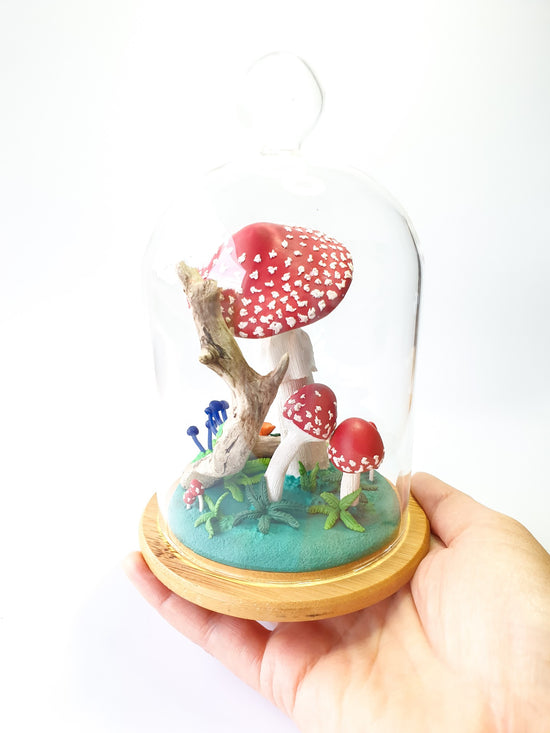 A glass cloche is held in a hand and inside it is a mushroom and forest scene with a log with tiny blue mushrooms and lots of red and white spotty amanita toadstool mushrooms