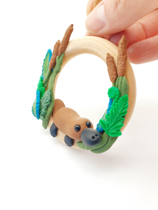 Polymer clay miniature platypus wreath sculpture. Hand made with incredible detail. Adorable, cute Australian native platypus animal wreath sculpture on a wooden timber wreath surrounded by sculpted ferns and plants