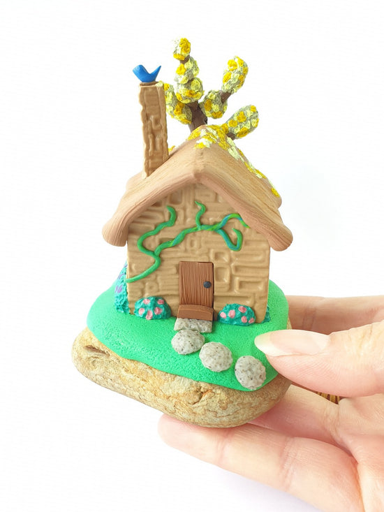 A cute miniature cottage sculpted out of clay suits on a rock in the artists hand. It has a wattle tree out the back and a tiny blue wren on the chimney.